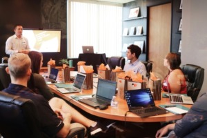 Meeting room with one person presenting and others sitting around a table with laptops in front of them